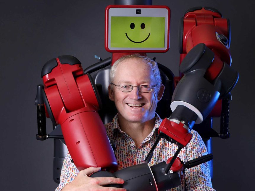 Baxter the robot wraps his arms around Toby Walsh, who is sitting on a chair in front of it