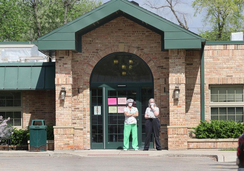 Two people talk outside the front entrance of Pine Acres Rehab and Living Center in DeKalb Thursday afternoon as signs posted on the door warn that there are confirmed cases of COVID-19 in the building. Nine staff members and 31 residents at the facility have tested positive for COVID-19. One resident has died as a result of complications from the disease.