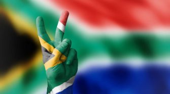 South African flag in the colours red, blue, black, green, gold and white in the background and painted on a person’s hand who is holding a peace sign.