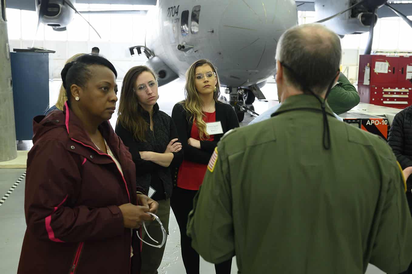 Students stand in front of a military plane listening to a man in a jumpsuit