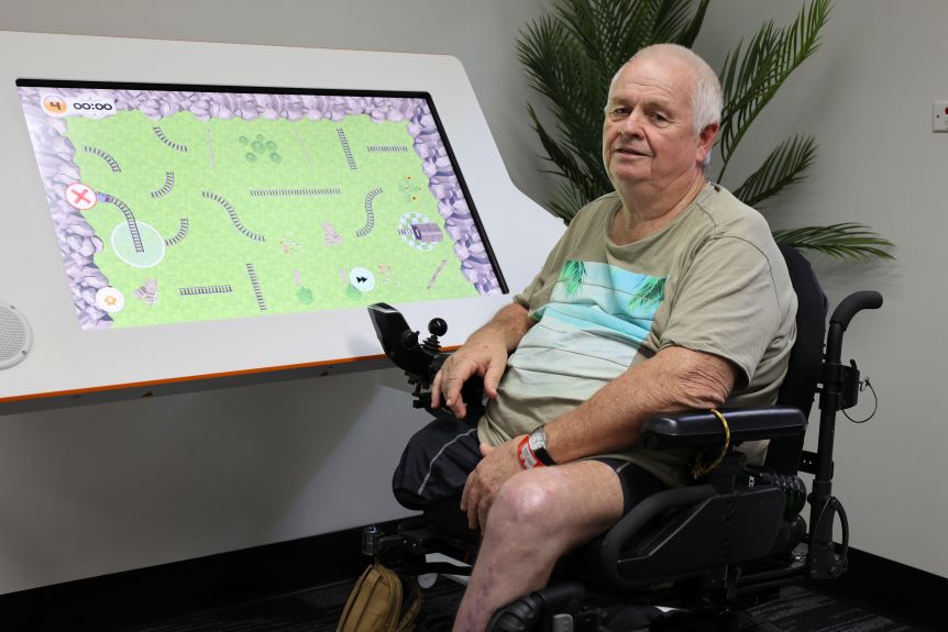A man in a wheelchair sitting in front of a large computer screen smiling at the camera