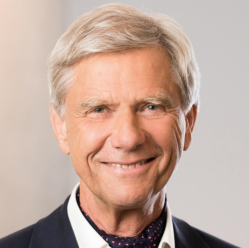 an older white man with white blond hair and a suit jacket smiling into camera