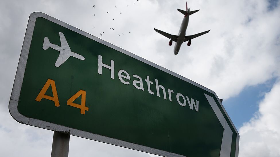 Plane flies over a road sign for Heathrow Airport