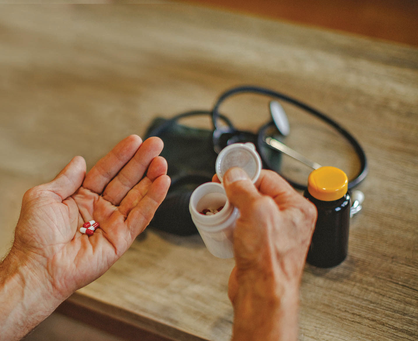 photo of the hands of an older man with acetaminophen capsules in the left hand and the medication bottle in the right, with a blood pressure cuff on a table in the background