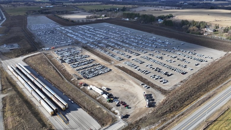 An aerial image shows dozens of cars parked on a dusty gravel lot. 