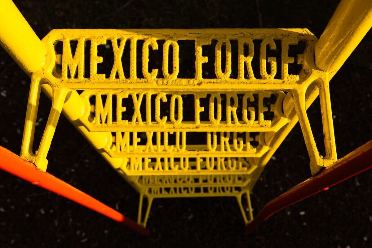 A yellow-painted metal playground ladder, viewed from the top. The steps say 'MEXICO FORGE'.